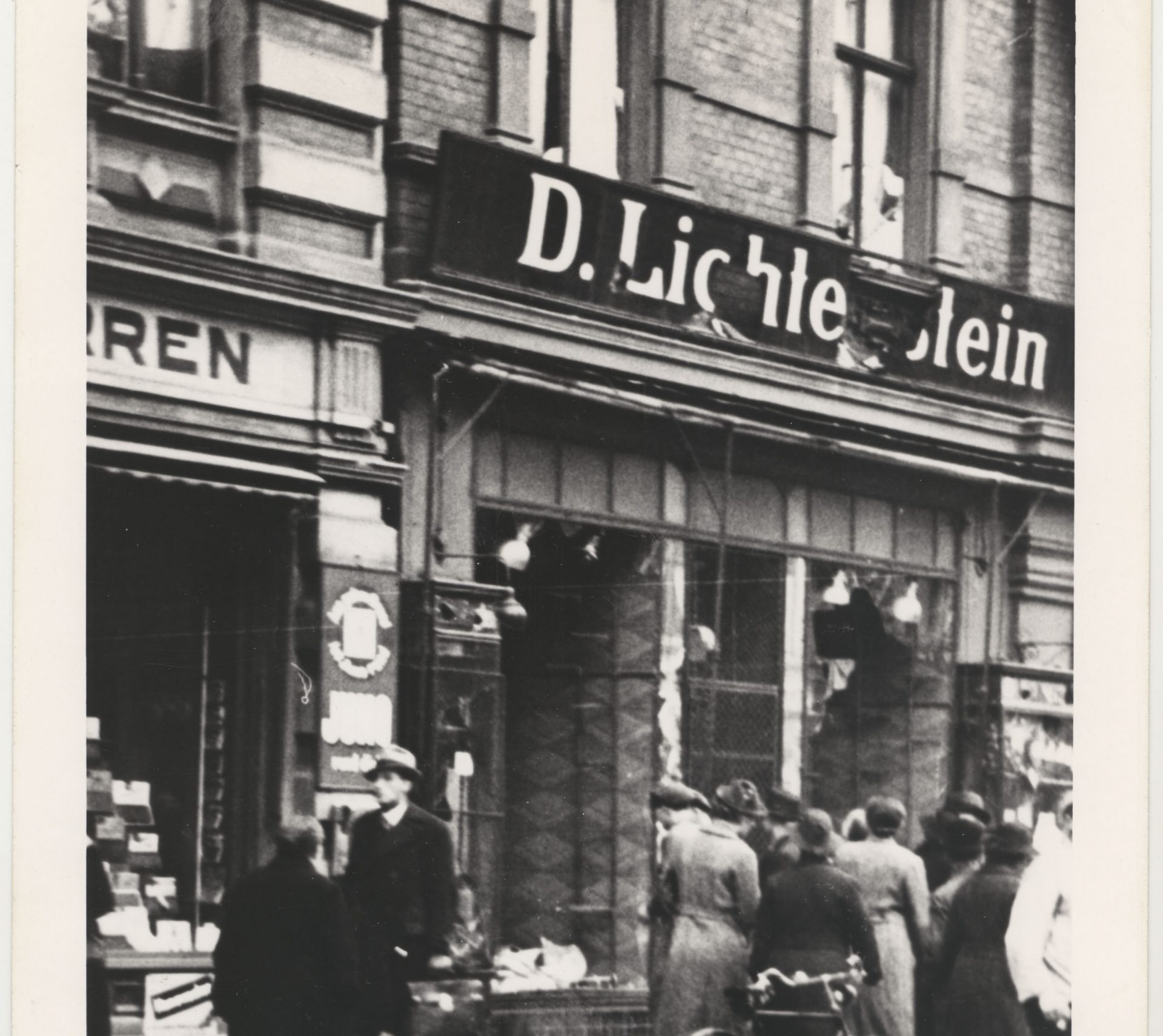 Cette photographie montre une devanture de magasin juif vandalisée au cours de Kristallnacht. Des passants regardent à l’intérieur d’une vitrine brisée sous l’écriteau ‘’D. Lichtenstein’’.