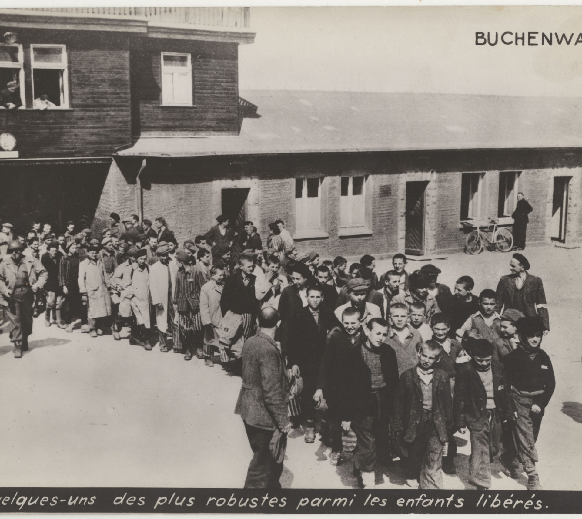 Photograph of a group of children who have been liberated and are escorted out of Buchenwald’s main gate by the American army. Stanley Weinstein is in front of the fourth row and wears a hat which he made himself. 